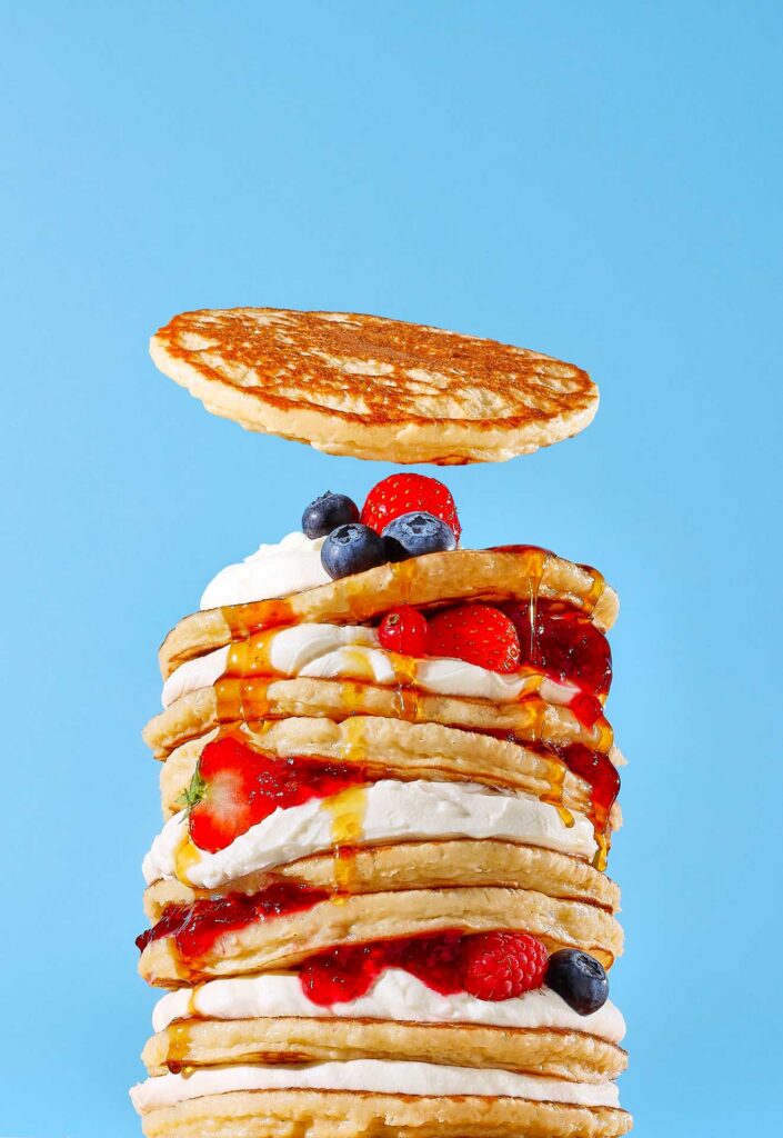 Stack of pancakes layered with whipped cream, berries, jam, and syrup, with one pancake hovering above the stack.
