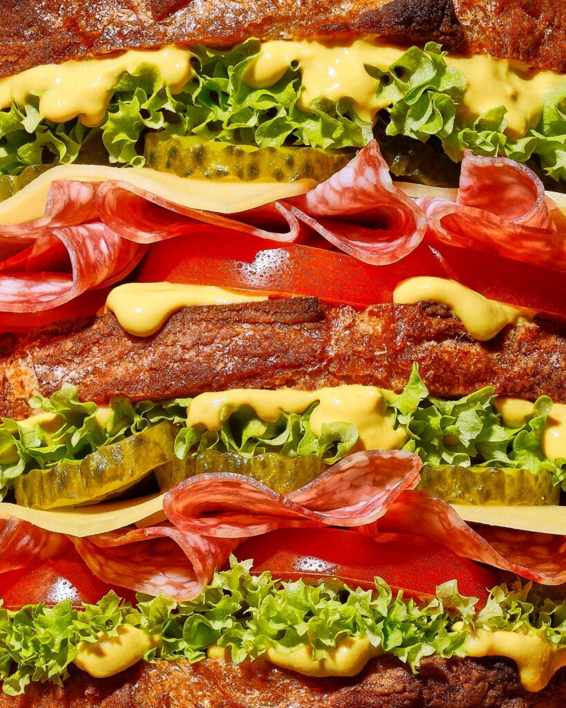 Extreme close-up of layered sandwich ingredients including fried chicken, lettuce, pickles, cheese, tomato, and sauce.