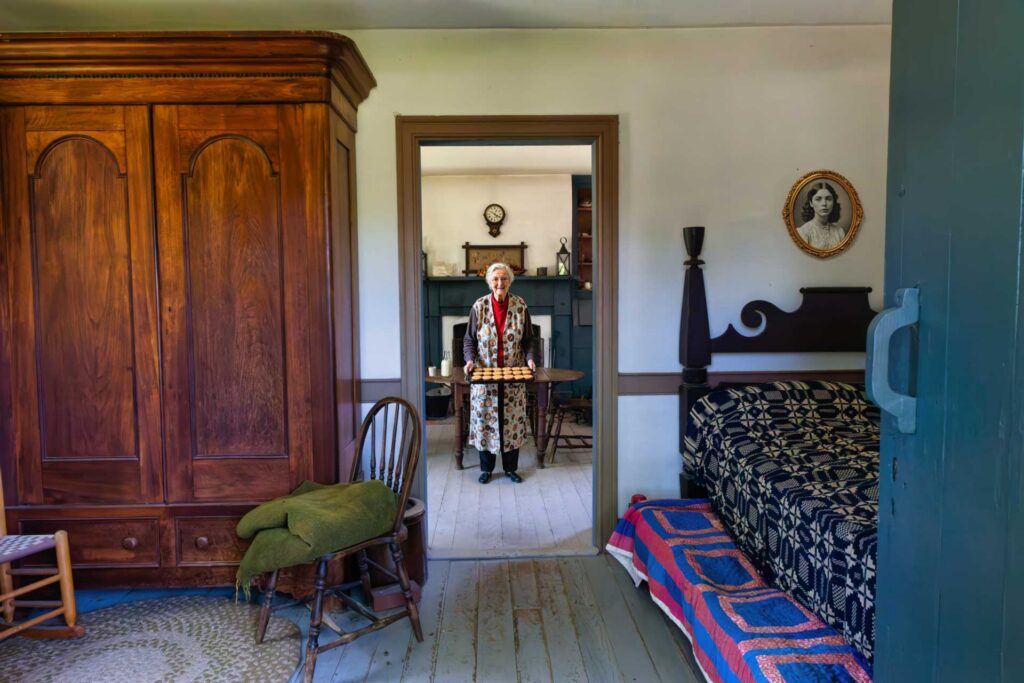 Historic bedroom with a wooden wardrobe and quilted bed, viewed through a doorway where an elderly woman stands holding a tray.