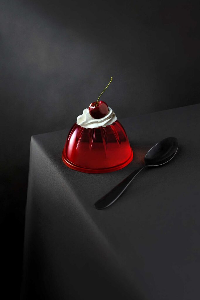 Red gelatin dessert topped with whipped cream and a cherry on a dark tabletop beside a spoon.