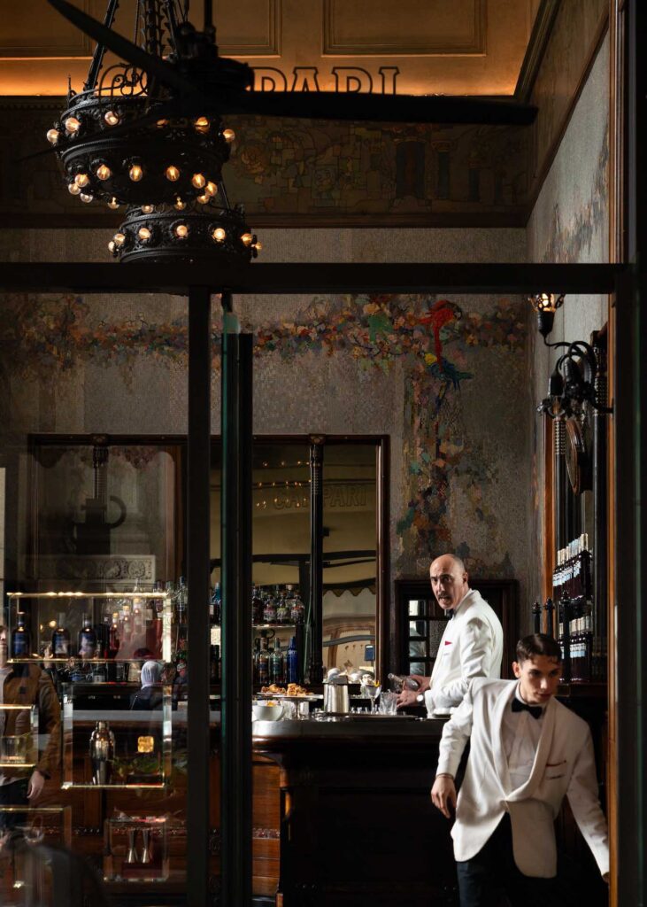 Elegant café or bar interior with ornate chandeliers, mural-covered walls, and two staff members in white jackets behind the counter.