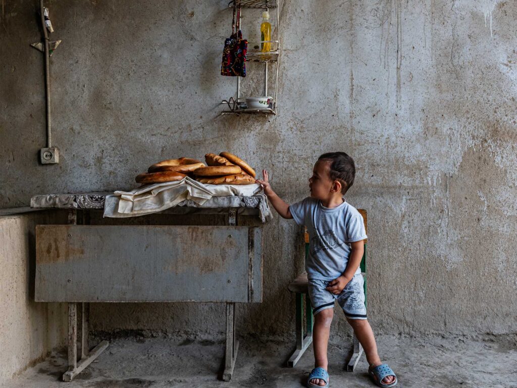 Young child reaching toward a stack of round loaves on a table inside a sparse room with worn concrete walls.