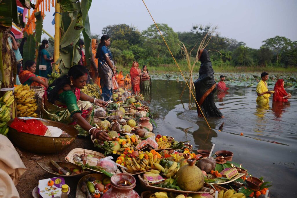 People standing and wading in a river during a religious offering ceremony, surrounded by fruit, flowers, and lit lamps.