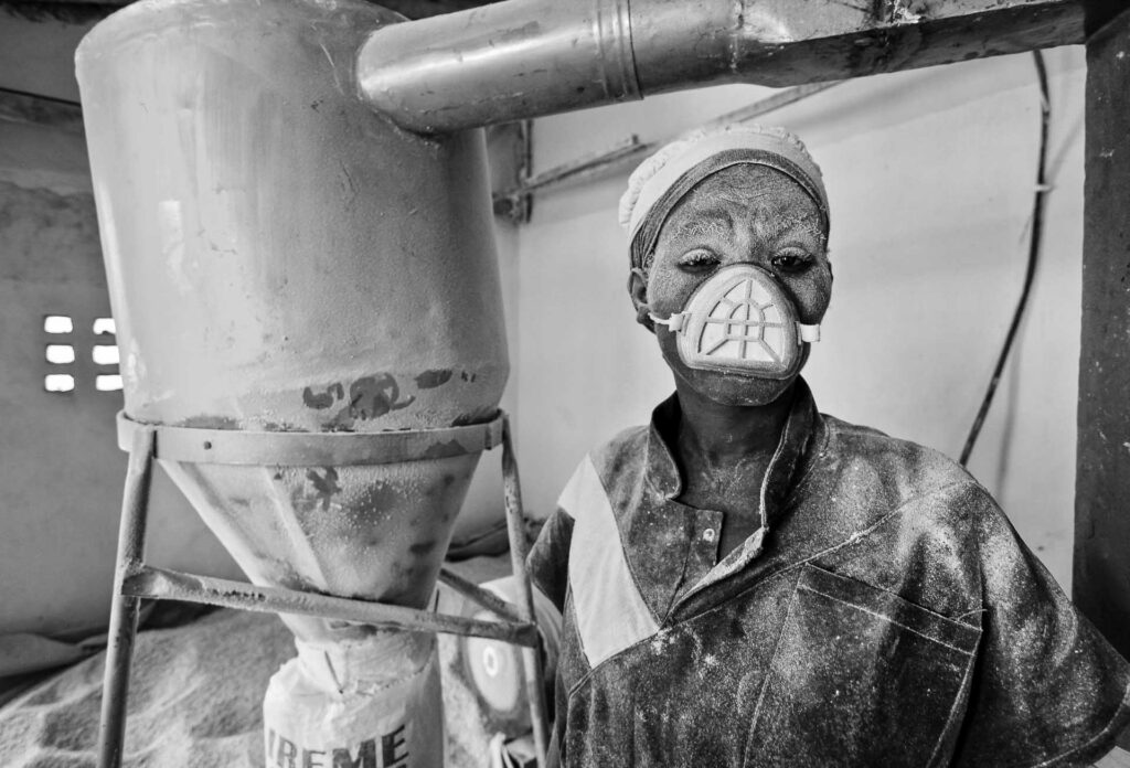 Worker covered in white dust wearing a protective mask beside industrial milling equipment.