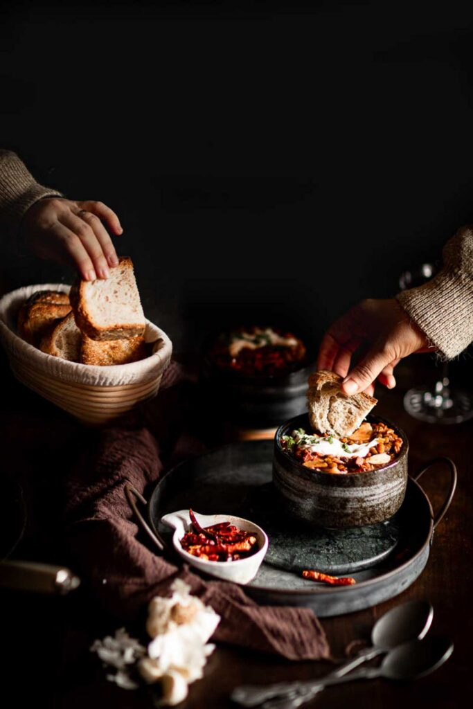 Hands dipping bread into a bowl of bean stew on a dimly lit table.