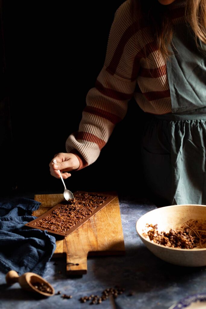 Person spooning chocolate mixture into a mold on a wooden board under dramatic side lighting.
