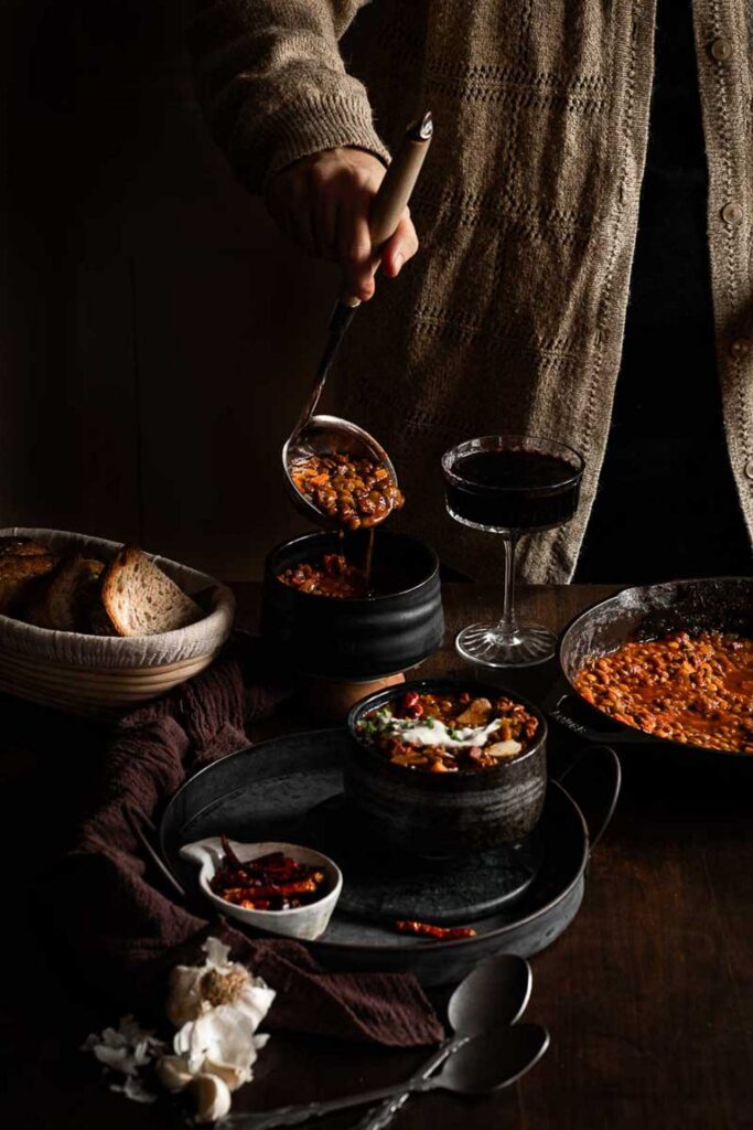Ladle pouring bean stew into a black bowl beside bread, a glass of red wine, and a skillet of stew.