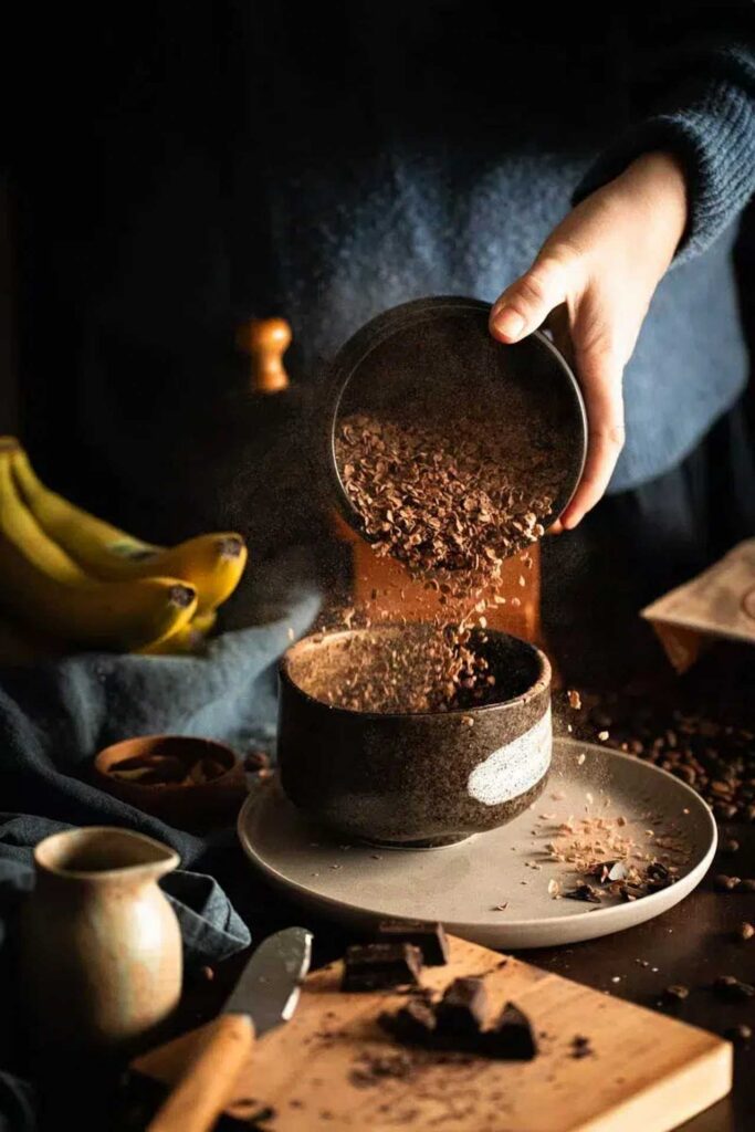 Hand sprinkling cacao nibs into a bowl, with bananas and chocolate pieces on a rustic tabletop.