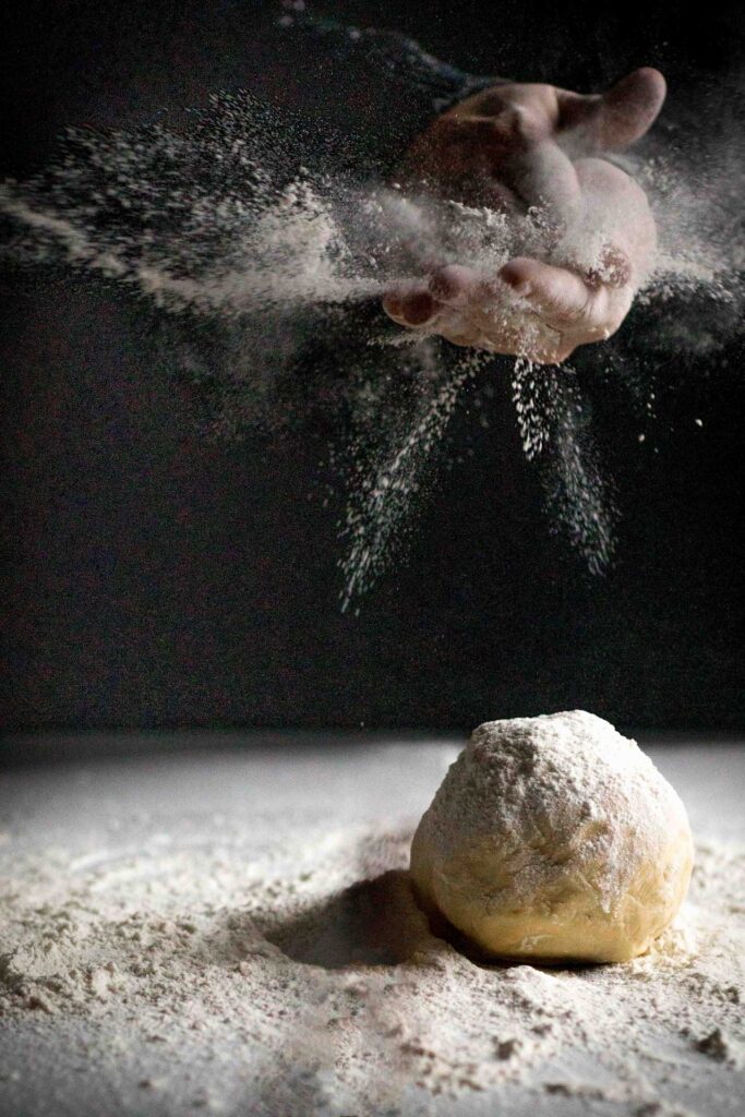 Flour bursting from cupped hands above a floured ball of dough on a work surface
