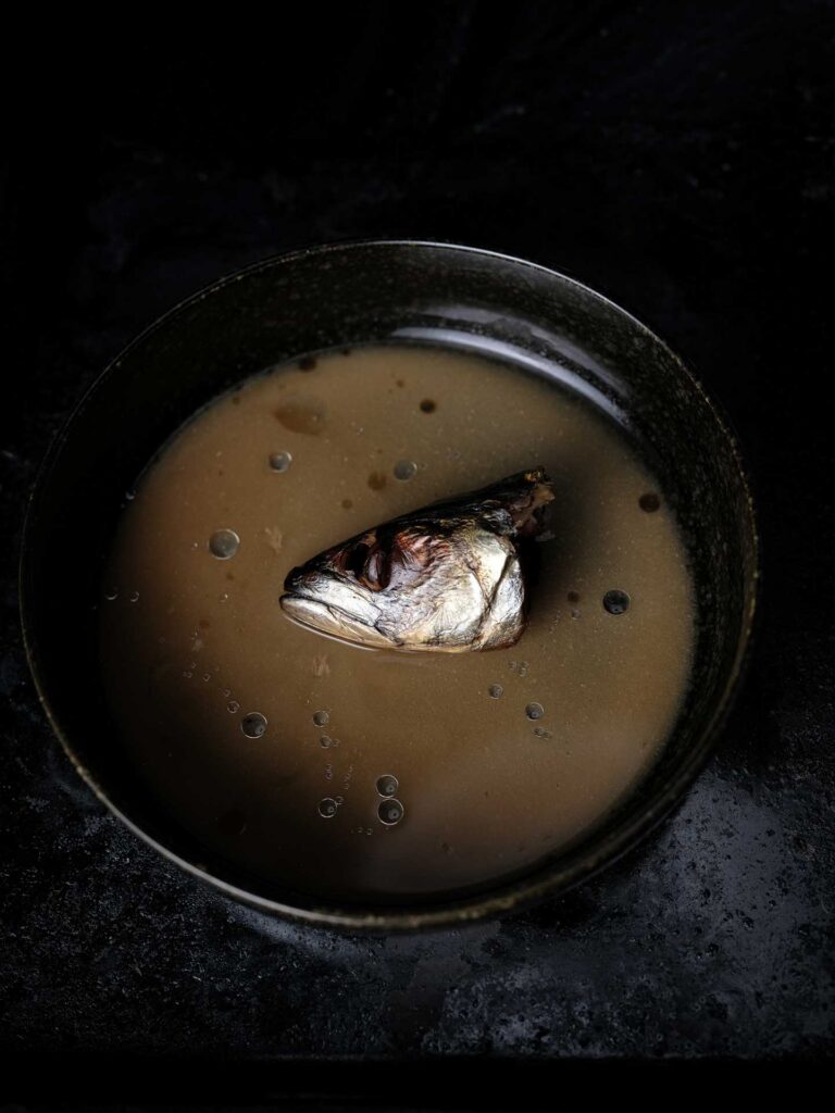 Fish head floating in a dark bowl of broth against a black background.