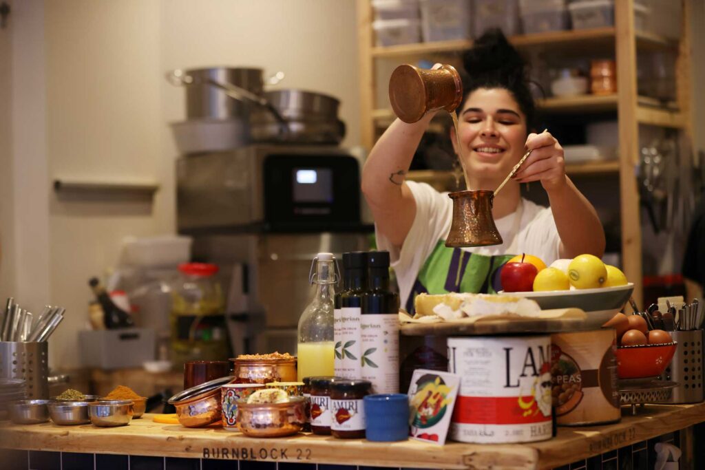 Cook pouring coffee from a copper pot into a small cezve in a home kitchen crowded with ingredients and cookware.