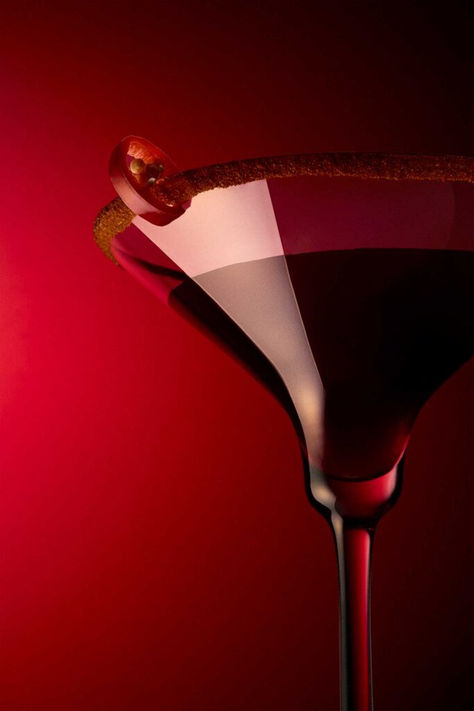 Martini glass with a chili-rim garnish and pepper slice against a vivid red background.