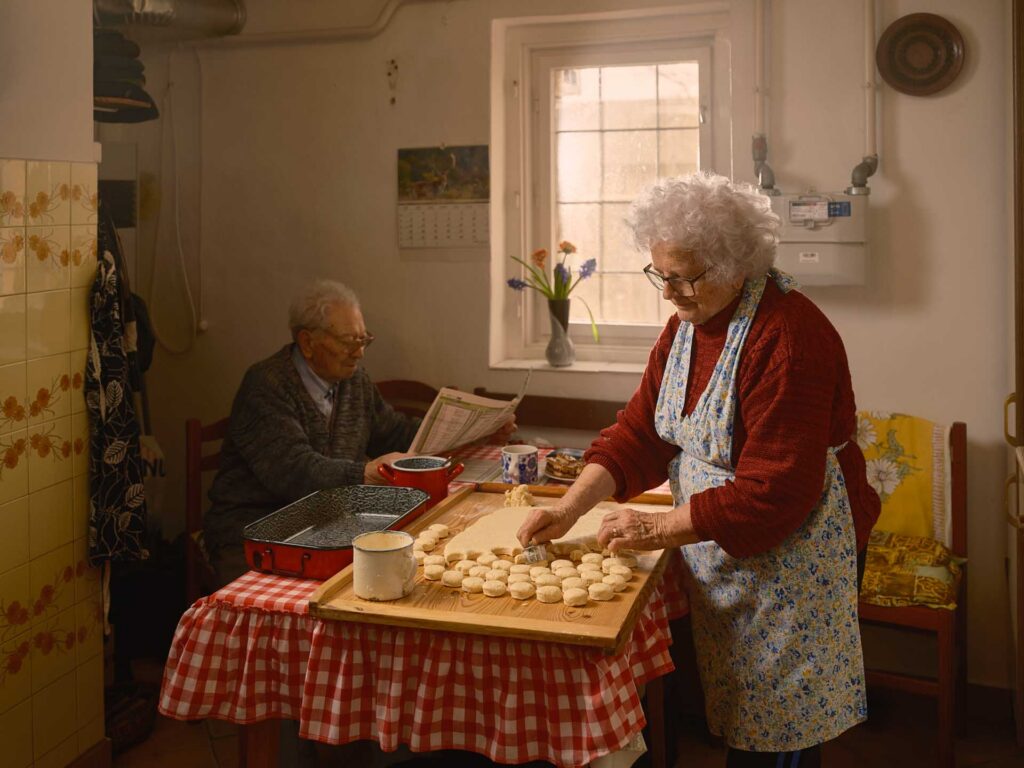 Elderly woman cutting dough into rounds at a kitchen table while an elderly man reads a newspaper nearby.