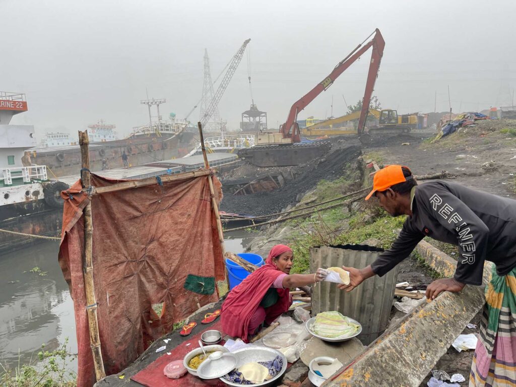 Woman handing flatbread to a man beside a canal in an industrial riverside area with cranes and boats in the background.