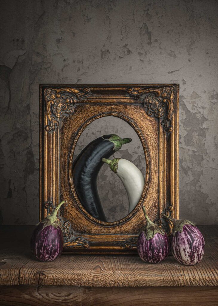 Black and white eggplants framed inside an ornate vintage picture frame, with striped eggplants arranged below.