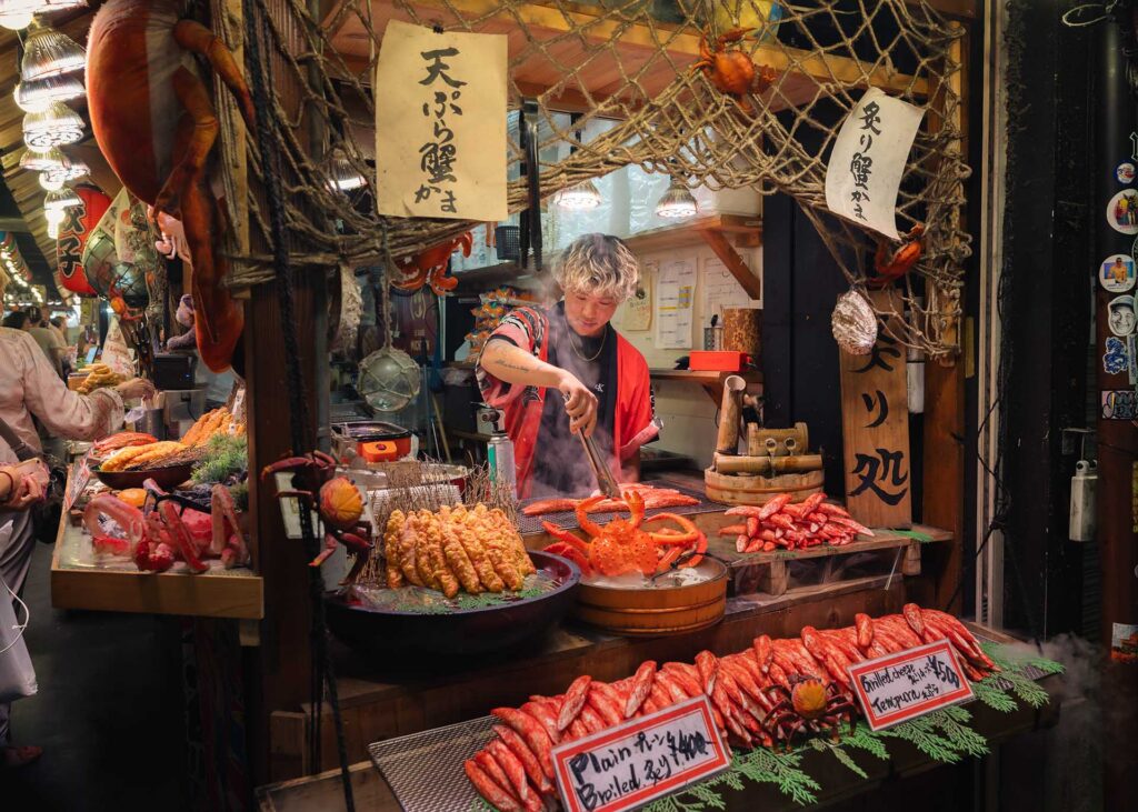 Vendor grilling crab legs at a seafood market stall decorated with fishing nets, crabs, and handwritten Japanese signs.