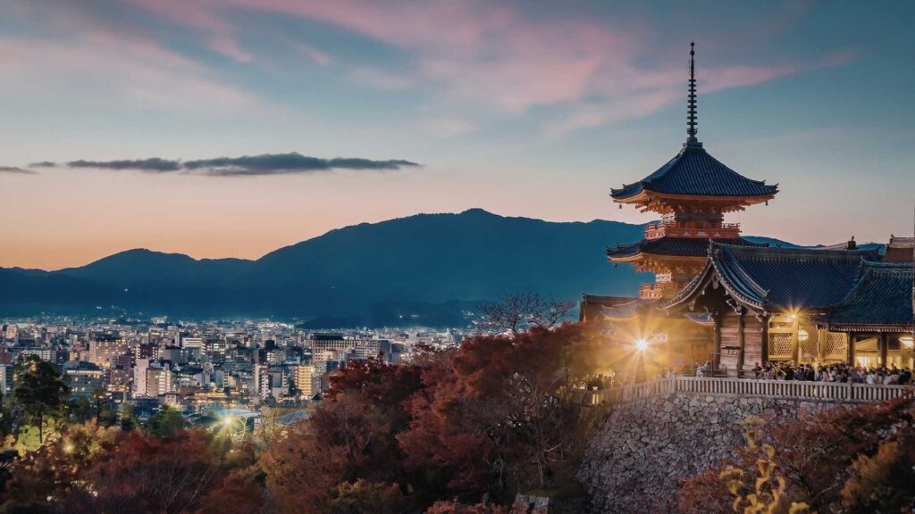 Kiyomizu-dera Temple overlooking Kyoto at twilight, with city lights and mountains in the distance.