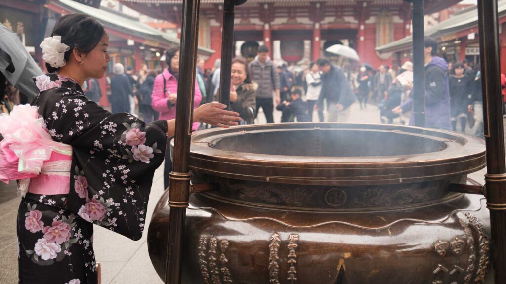 Woman in a floral kimono warming her hands over a large incense burner at a busy temple courtyard.