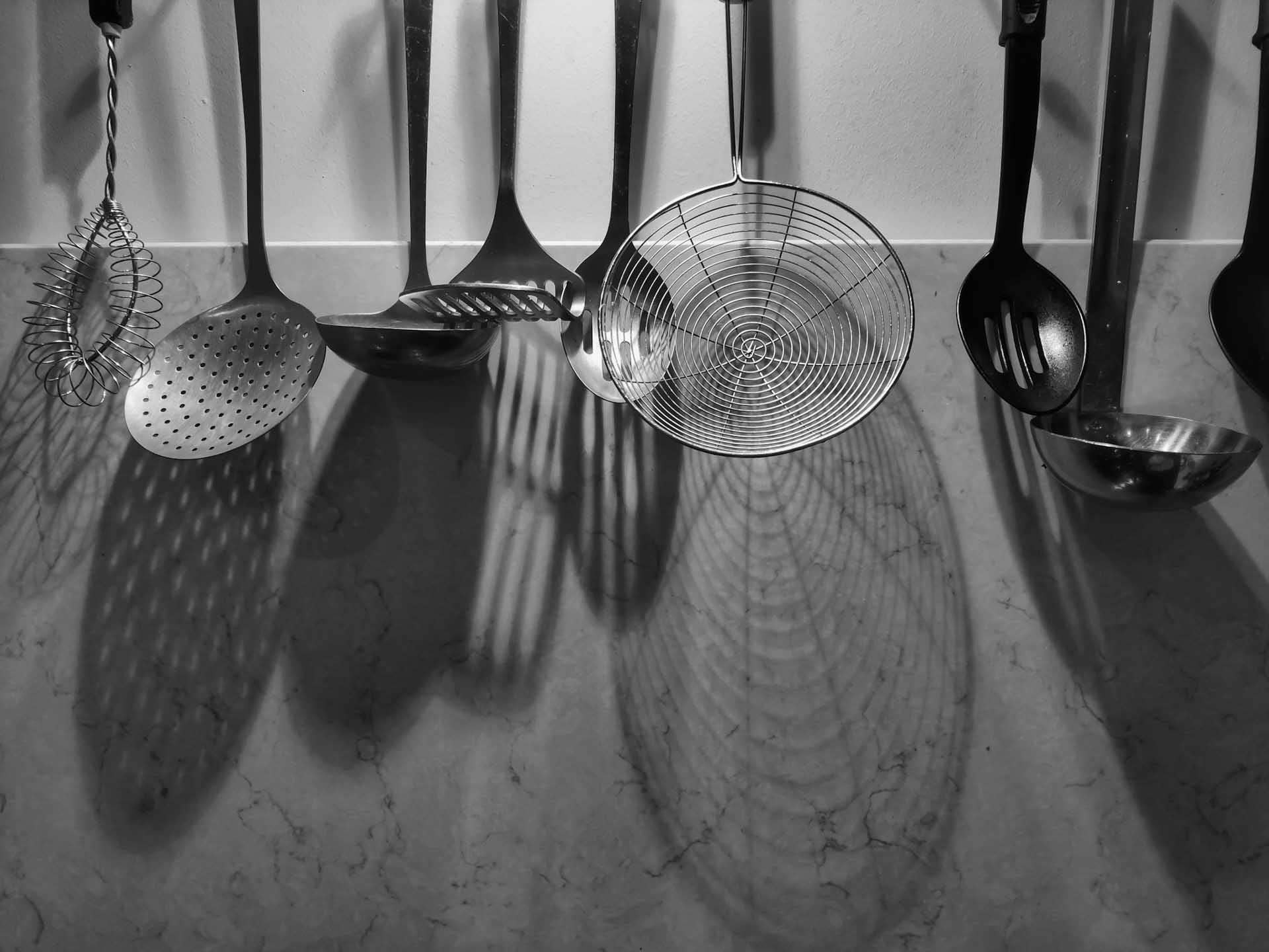 Black and white image of hanging kitchen utensils with patterned shadows falling across a marble splashback.