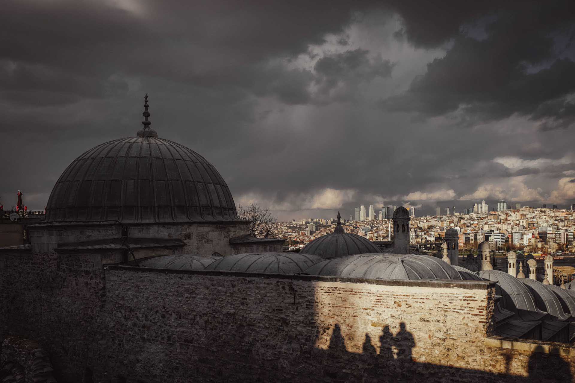 Storm clouds over Istanbul rooftops with silhouettes of people cast onto a sunlit wall.