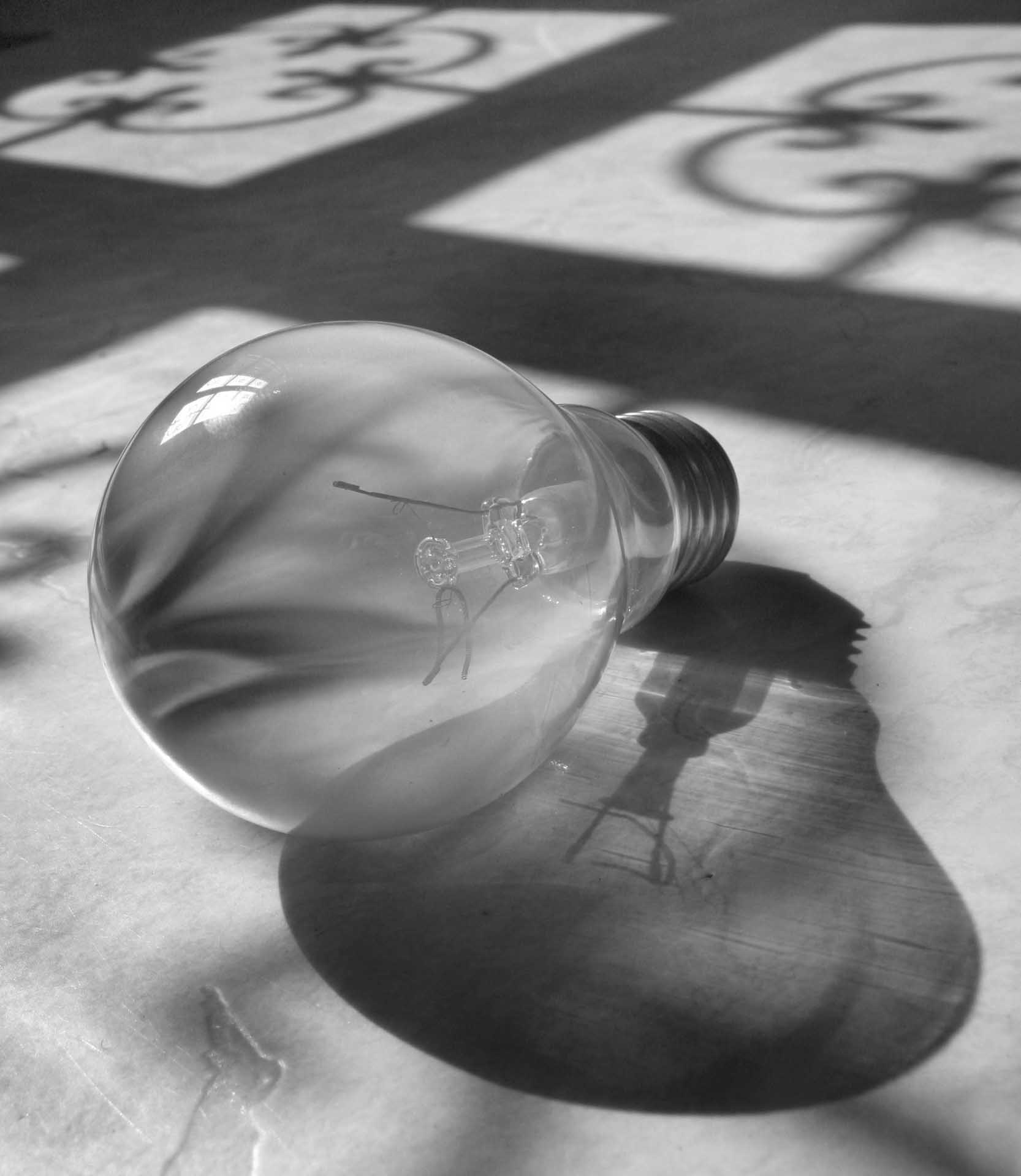 Black and white photograph of a light bulb lying on a surface with a large shadow and window patterns behind.