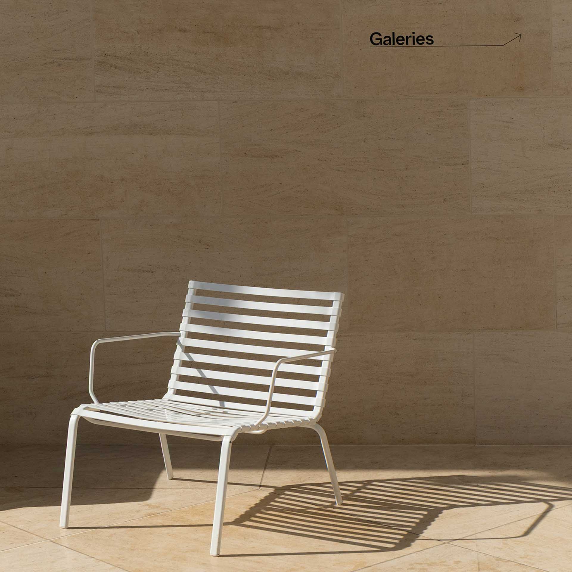 White metal chair casting striped shadows against a stone wall marked with a gallery sign.