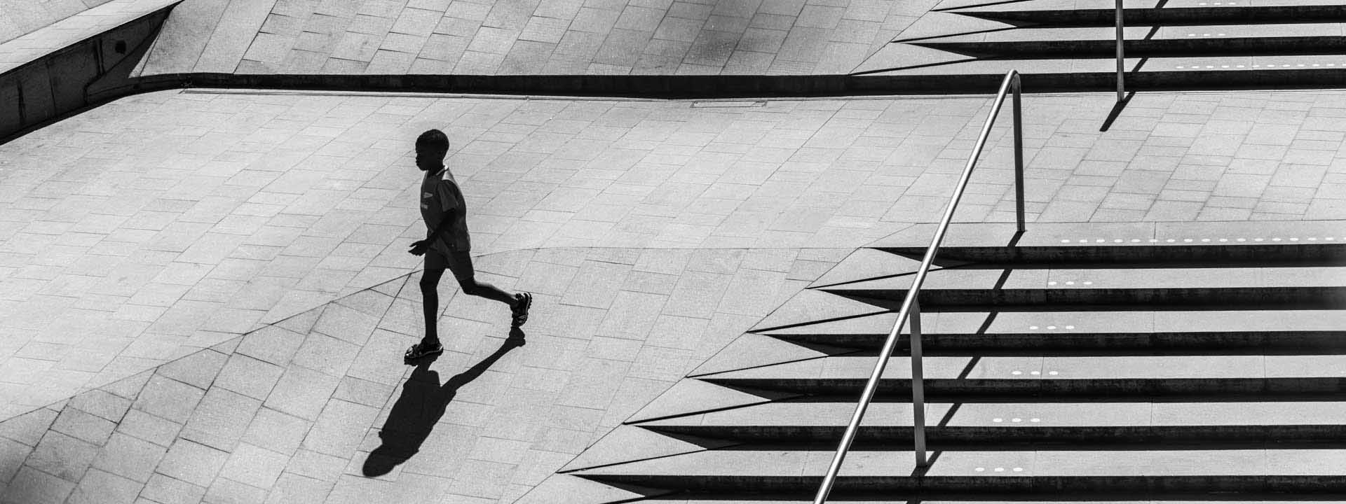 Black and white image of a child running across a plaza with geometric shadows from nearby steps.