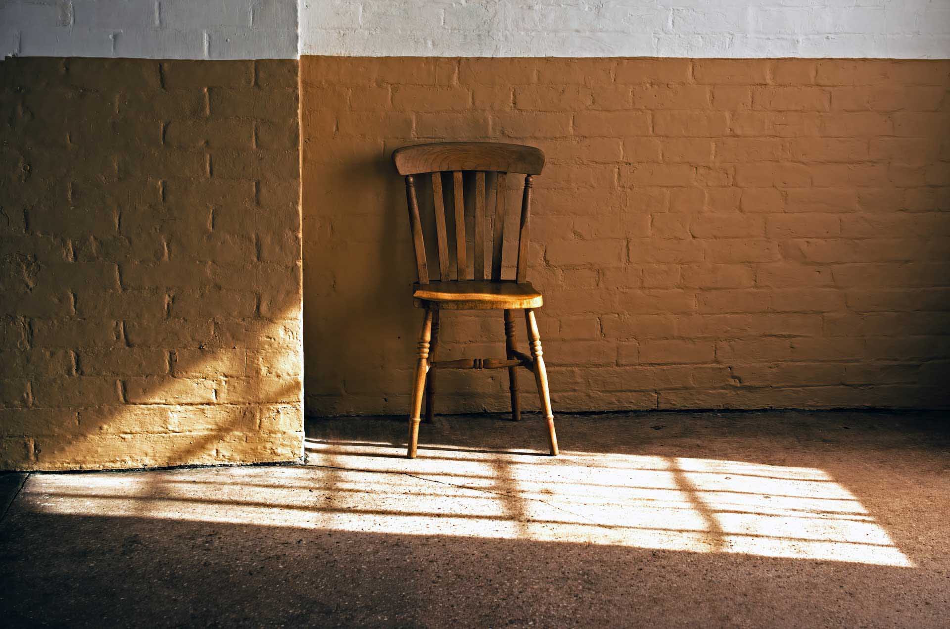 Single wooden chair illuminated by sunlight streaming through a window onto a brick wall.
