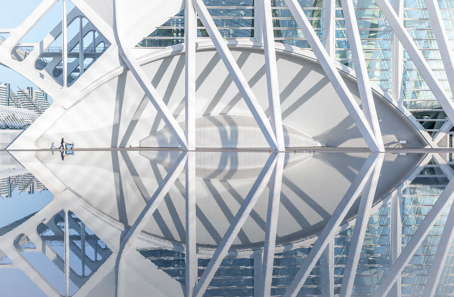 Modern white architecture reflected in still water with strong diagonal shadows and a tiny figure pushing a trolley.