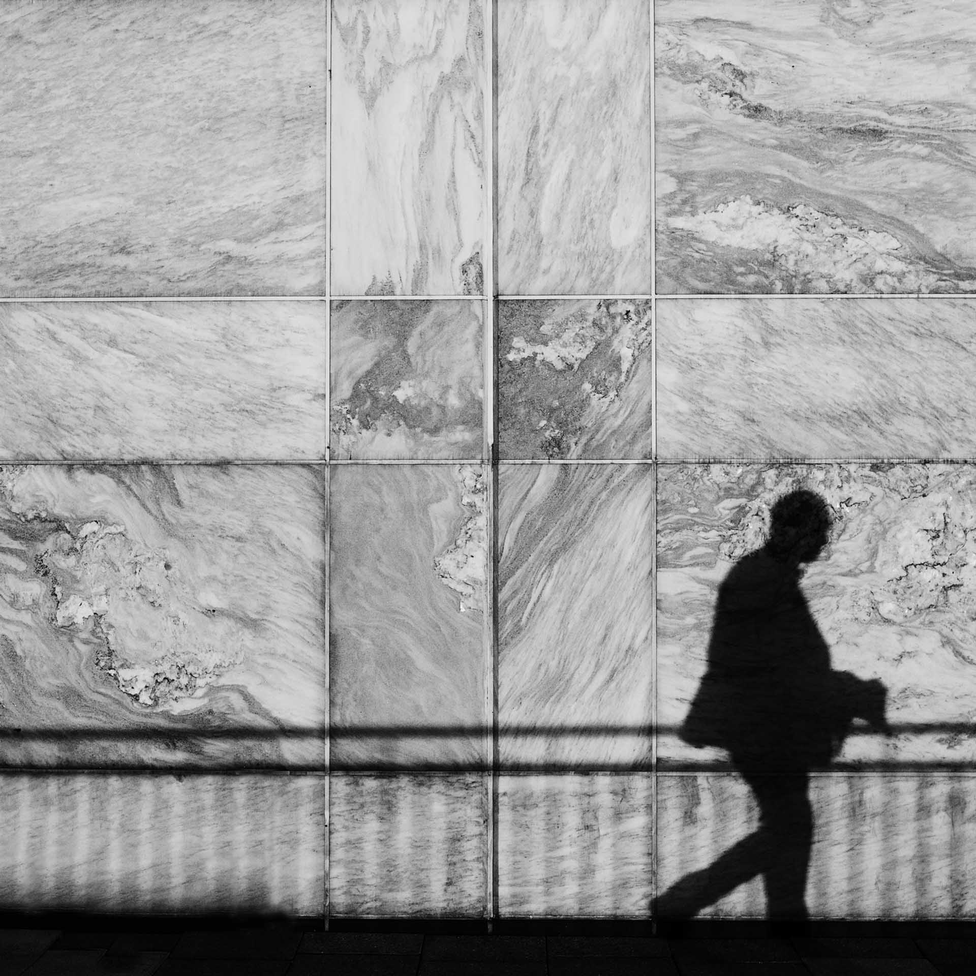 Silhouette of a person walking past a marble wall with railings casting shadows across the scene.