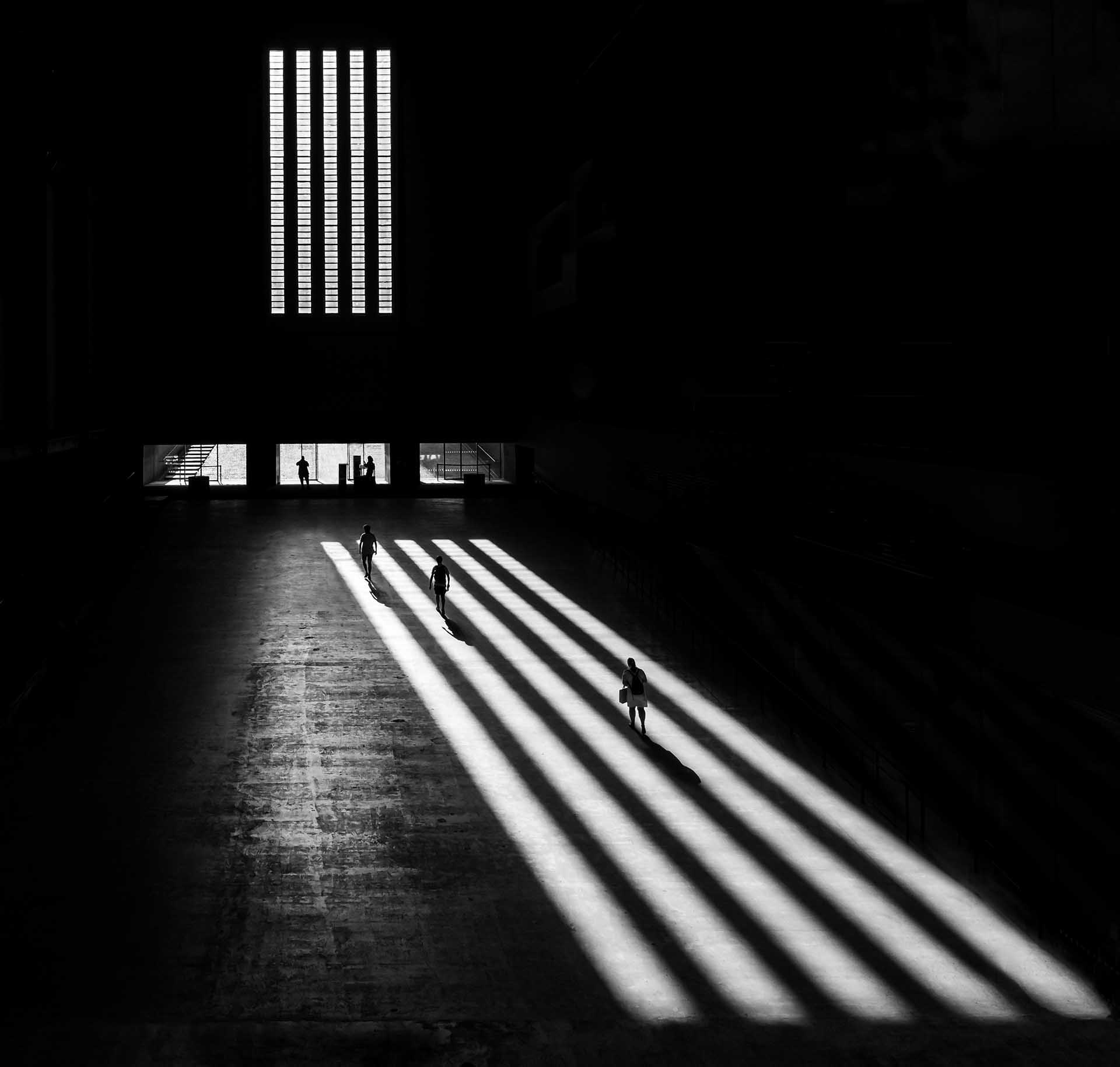 Three people walking through bands of sunlight and shadow inside a vast dark hall.