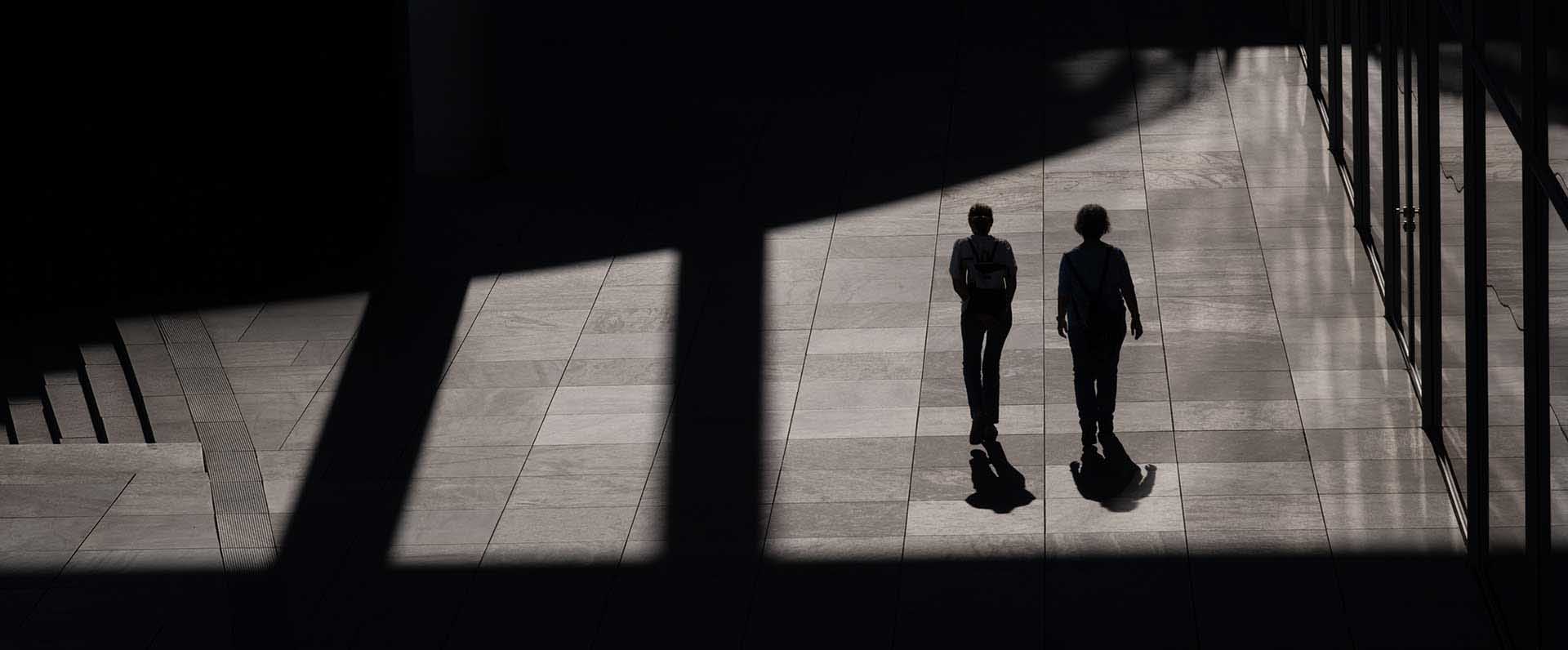 Two figures walking through dramatic shadows cast across a tiled indoor space.