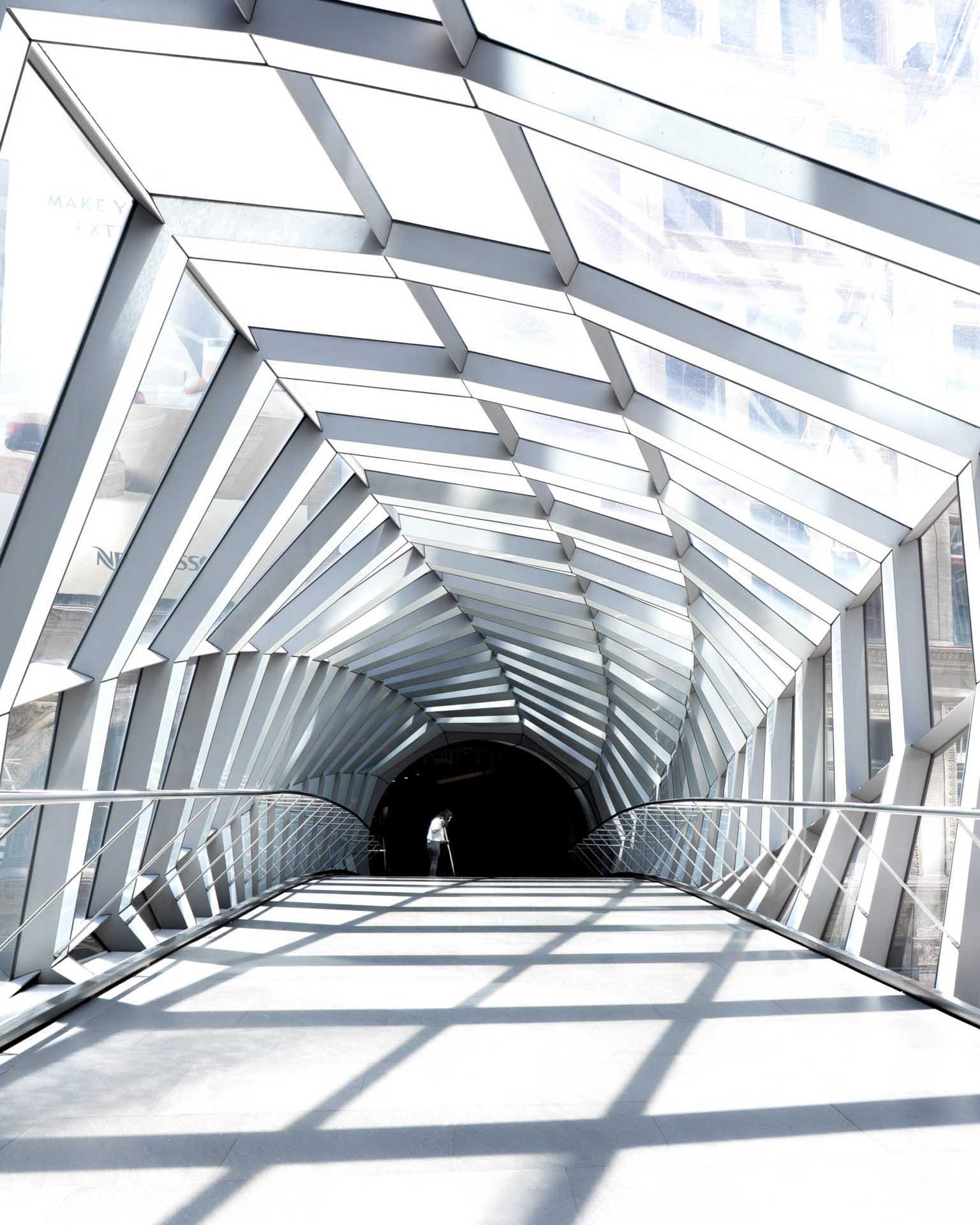 Geometric glass walkway with repeating shadows leading towards a dark opening and a lone figure.