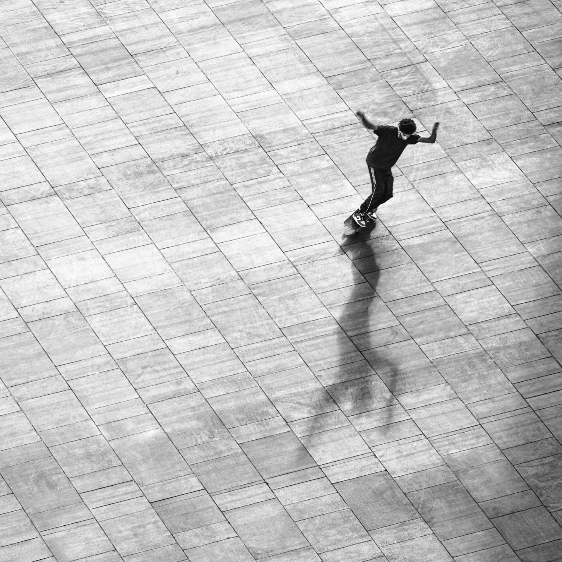 Black and white image of a skateboarder casting a long shadow across a paved square.