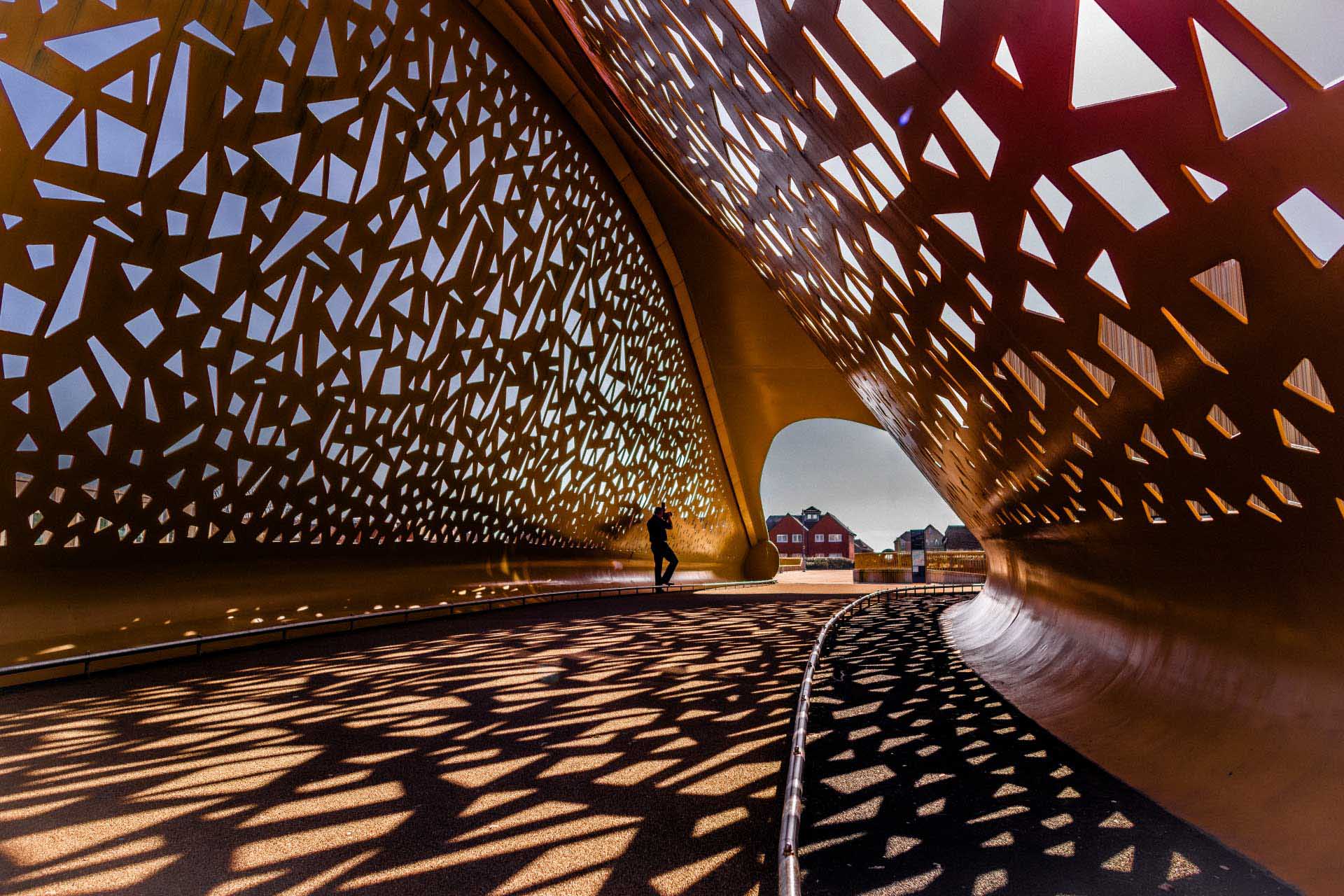 Golden perforated structure casting intricate shadows across a walkway with a silhouetted figure.