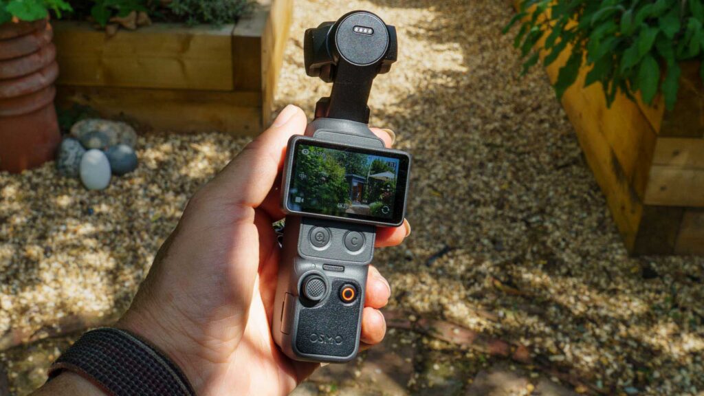 Hand holding a DJI Osmo Pocket 4 outdoors; the touchscreen displays a garden scene being recorded.