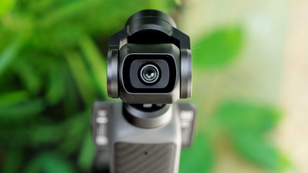 Detailed close-up of the DJI Osmo Pocket 4 camera lens and gimbal assembly with green foliage blurred behind it.