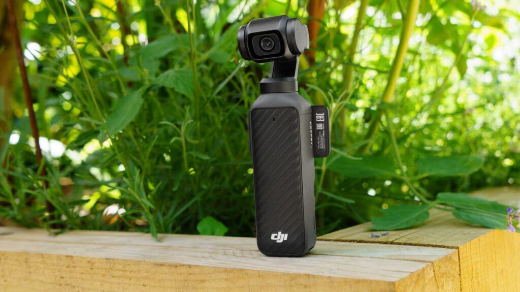 DJI Osmo Pocket 4 standing on a wooden surface in a garden setting, surrounded by green plants.
