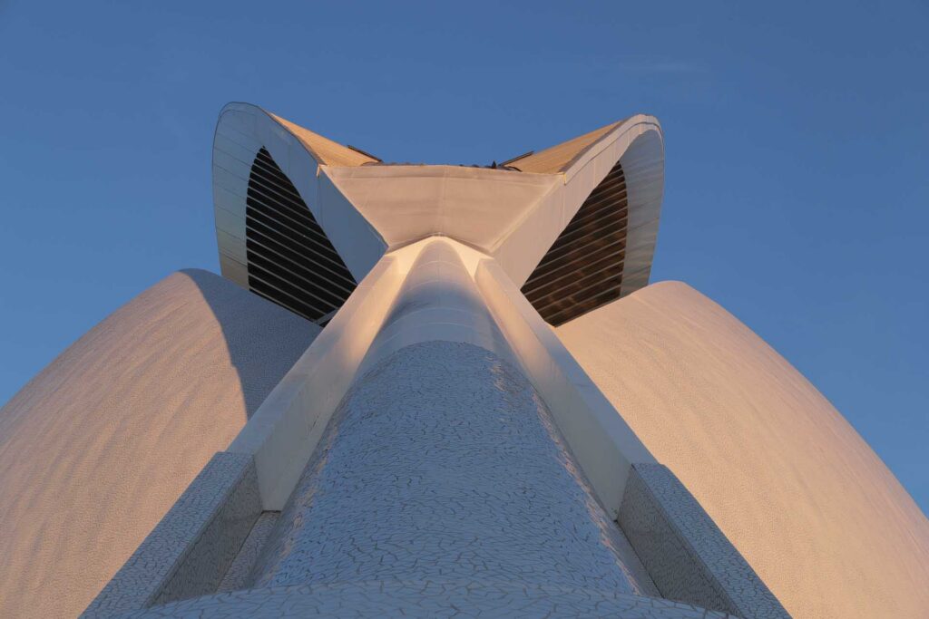 Low-angle view of a white futuristic structure with curved forms and dark louvered openings against a clear blue sky.