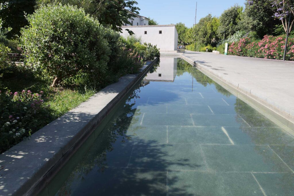 Long rectangular reflecting pool bordered by greenery, leading toward a white building at the end of a garden path.