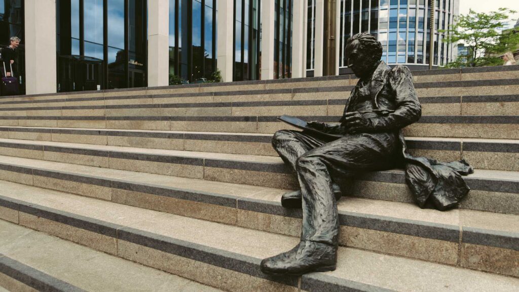 Bronze statue of a seated figure reading on wide stone steps outside a modern office building.