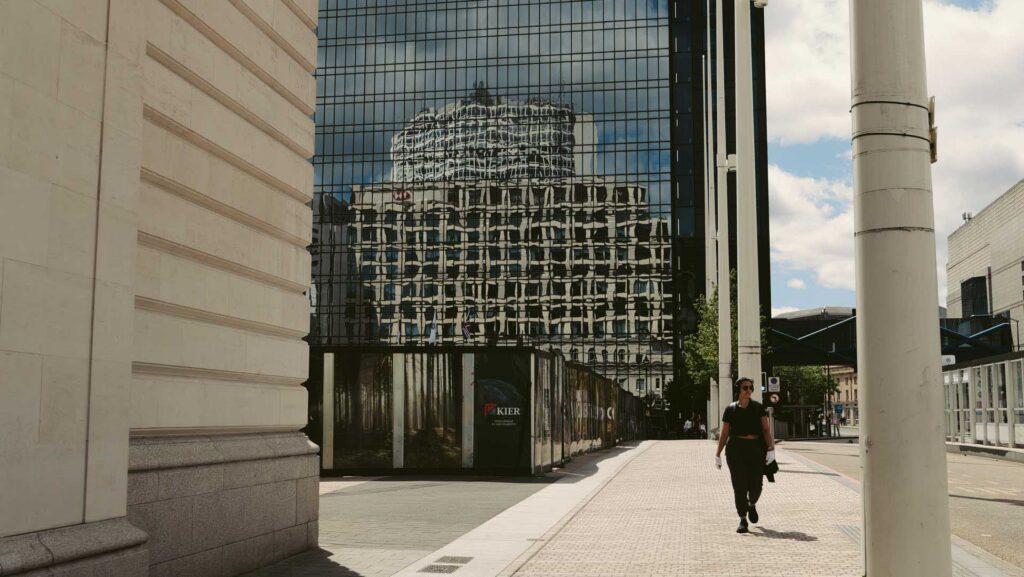 Glass office tower reflecting the distorted façade of a neighboring historic building, with a pedestrian walking past.