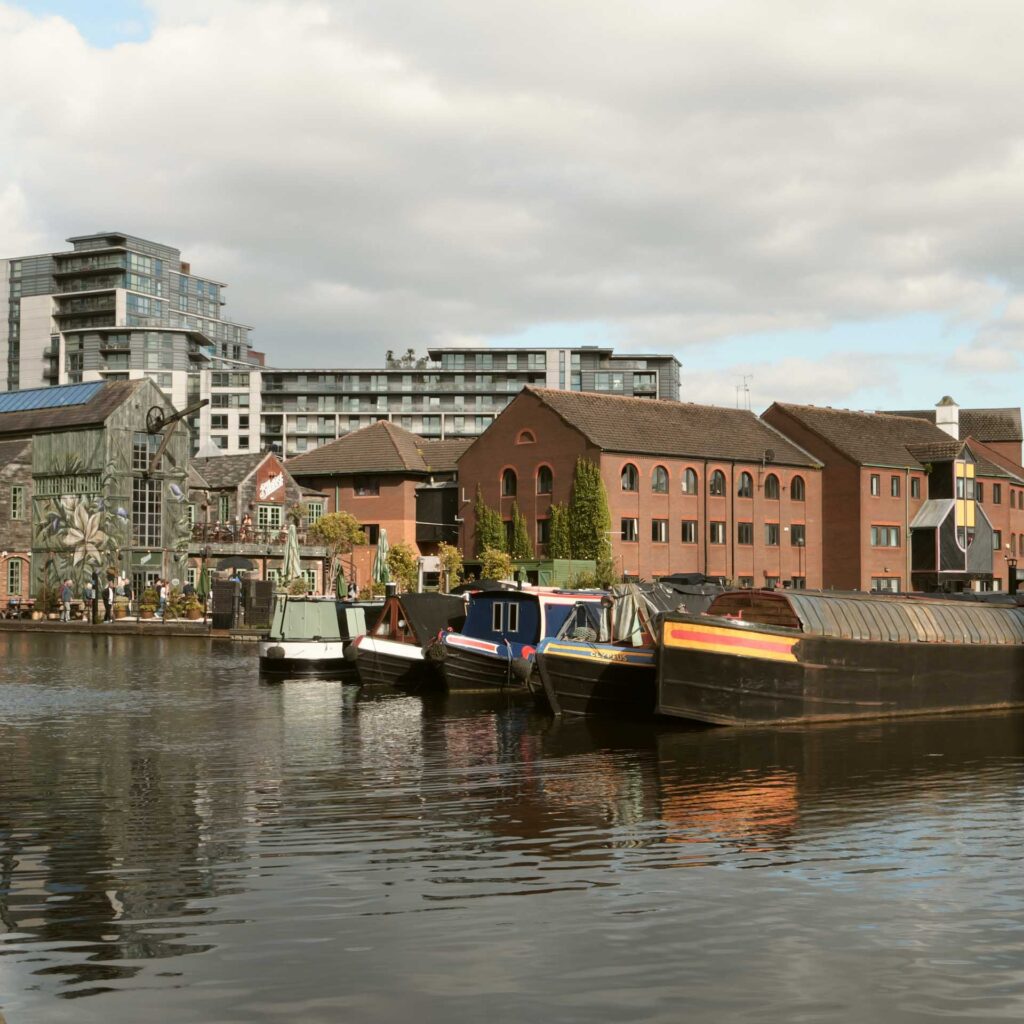 Waterside view of narrowboats and brick buildings under a cloudy sky, with modern apartment blocks behind.