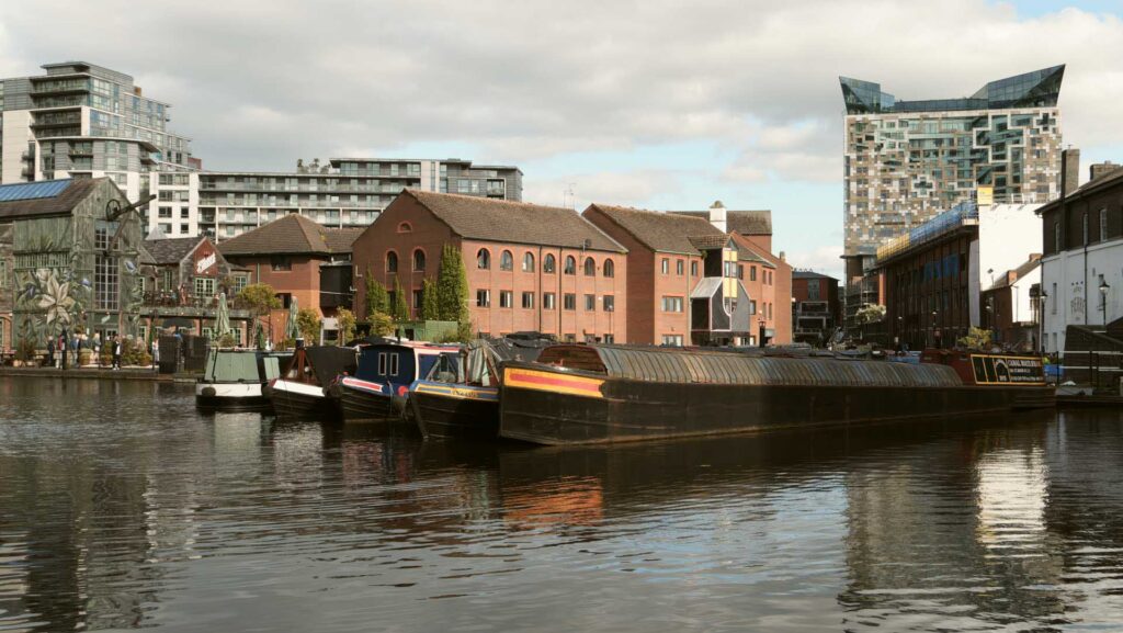 Waterside view of narrowboats and brick buildings under a cloudy sky, with modern apartment blocks behind.