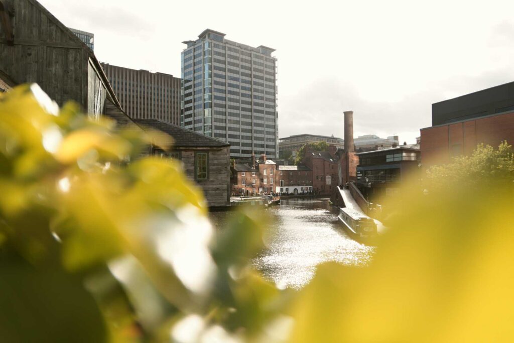 Sunlit canal scene framed by blurred yellow-green leaves, with historic industrial buildings and a bridge in the distance.