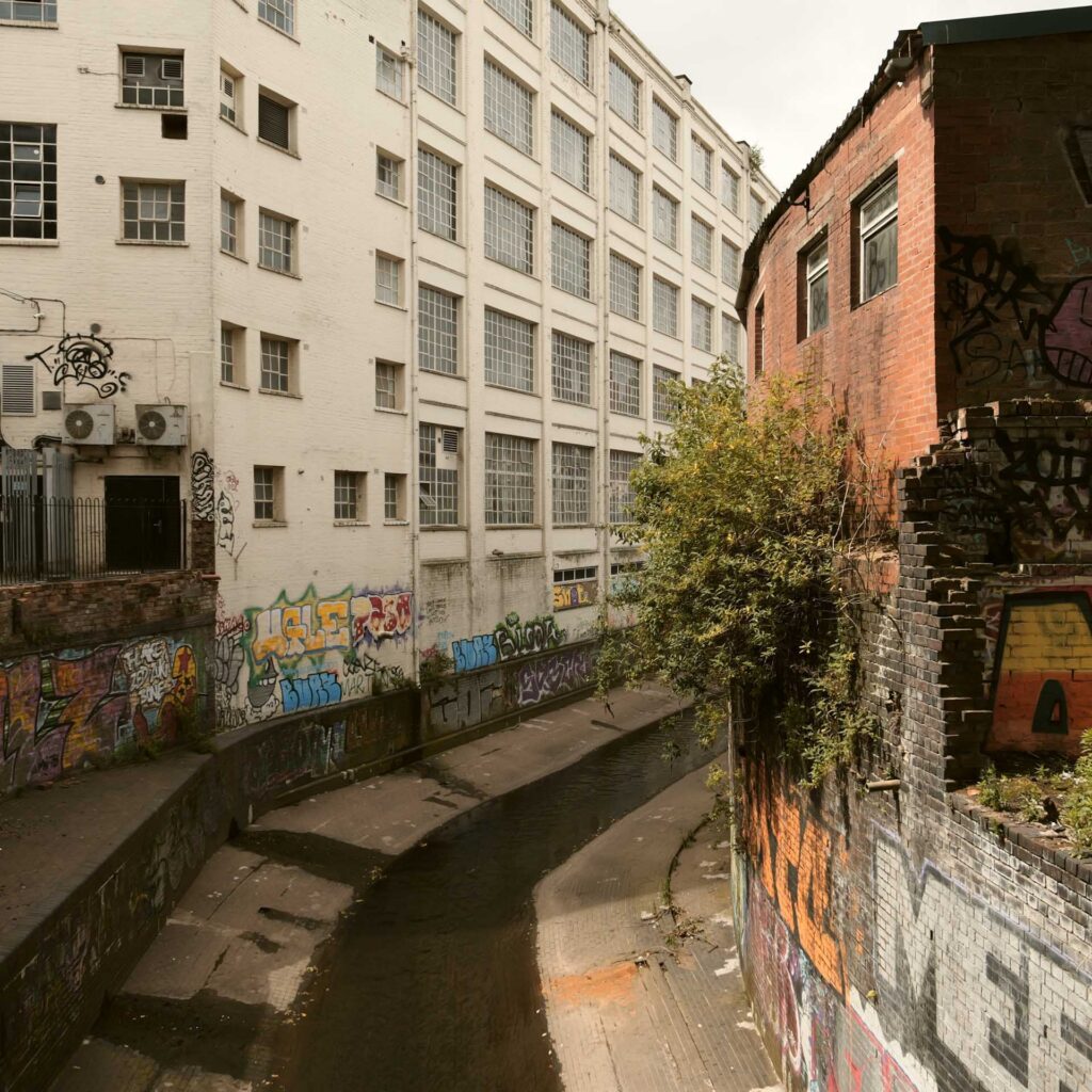 Narrow urban canal running between graffiti-covered brick and white industrial buildings.