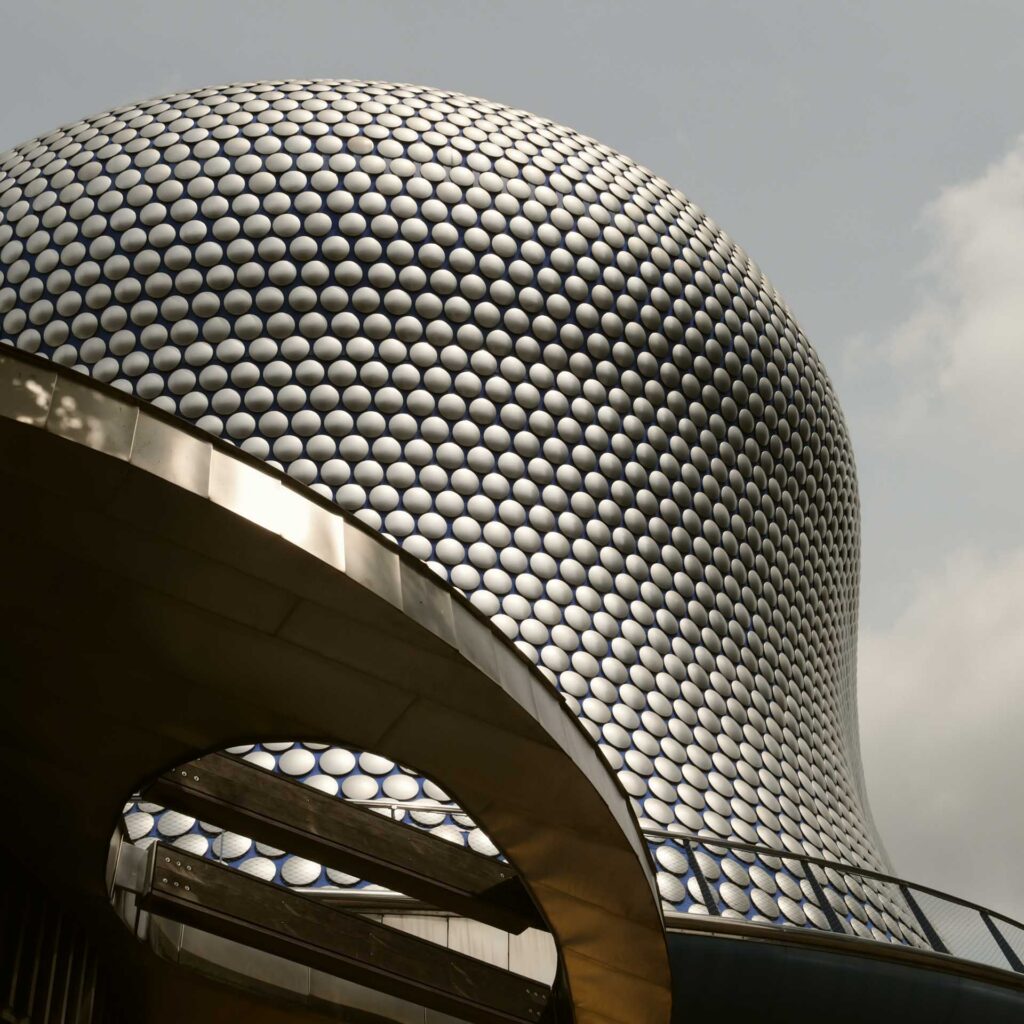 Curved exterior of a building covered in silver circular discs, viewed from below against a cloudy sky.
