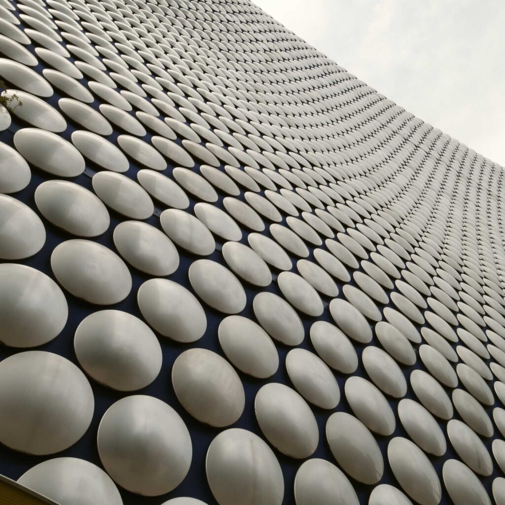 Sweeping upward view of a curved façade covered in silver circular panels receding toward the sky.