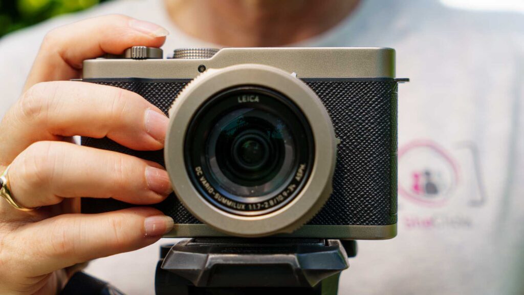 Close-up of Panasonic Lumix L10 front view mounted on a tripod, with a hand holding the camera body and the Leica-branded lens centered in frame.