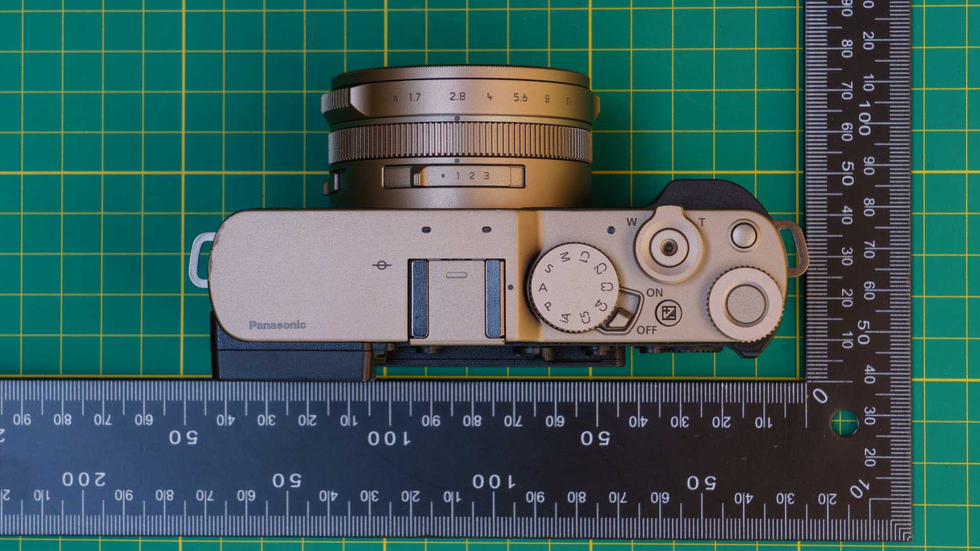 Top-down view of the Panasonic Lumix L10 placed on a cutting mat between measuring rulers, showing the camera’s controls and dimensions.